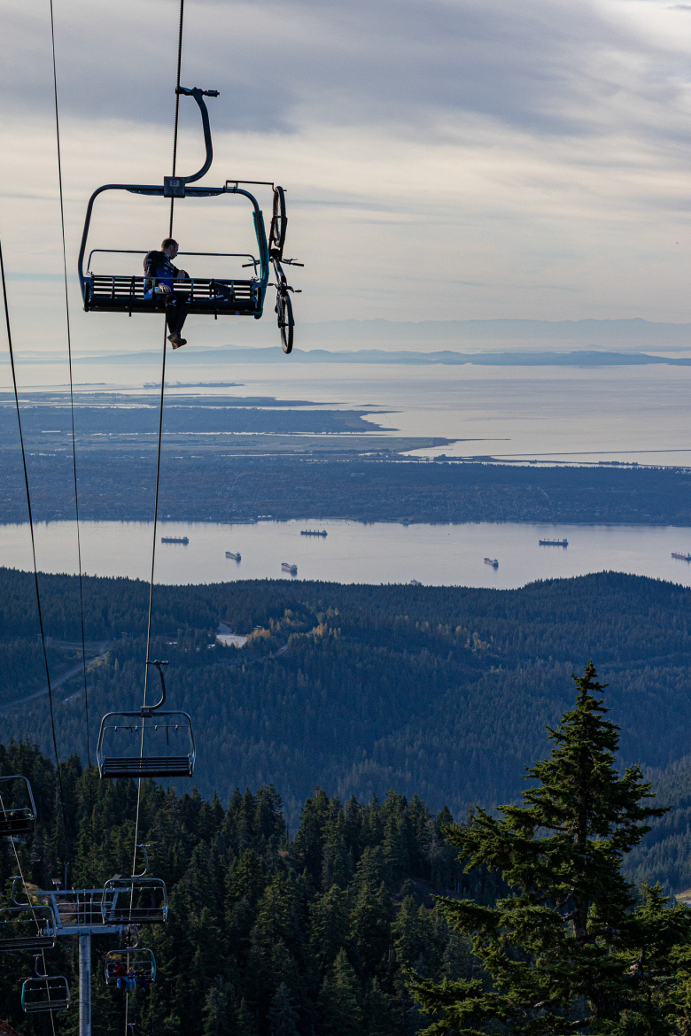 A person sitting on a chairlift with a bicycle attached, overlooking a vast landscape of trees and a bay with ships. The sky is partly cloudy, and mountains can be seen in the distance.