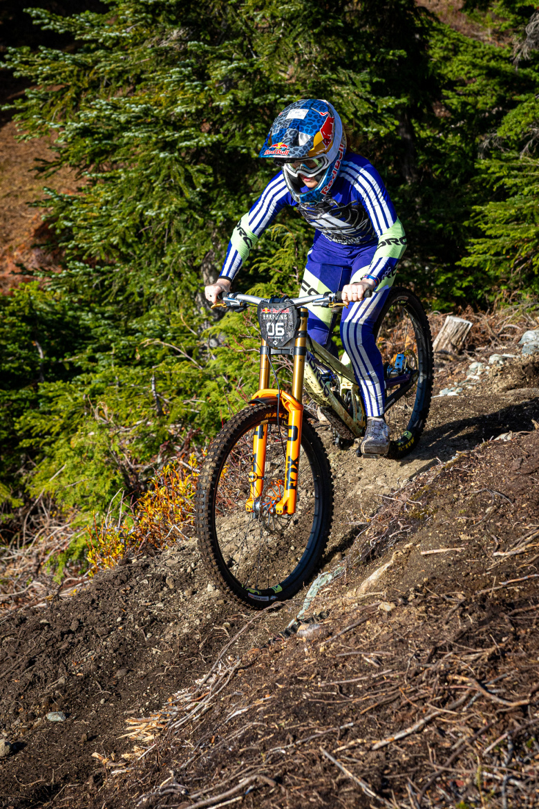 A mountain biker in protective gear rides on a dirt trail surrounded by trees and vegetation. The biker is leaning forward on a brightly colored bike, showcasing an intense focus as they navigate the terrain.