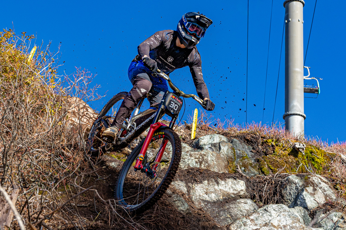 A mountain biker in protective gear is navigating a rocky downhill trail, kicking up dirt and debris as they descend. The background features blue skies and sparse vegetation, with a utility pole visible in the frame. The biker is wearing a helmet and a number plate.