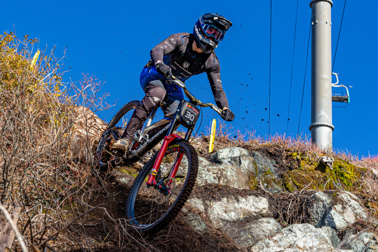 A mountain biker in protective gear is navigating a rocky downhill trail, kicking up dirt and debris as they descend. The background features blue skies and sparse vegetation, with a utility pole visible in the frame. The biker is wearing a helmet and a number plate.