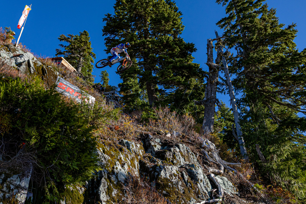 A mountain biker mid-air performing a jump off a rocky cliff, surrounded by lush green trees and a bright blue sky. A sponsor banner is visible on the rocky surface below.