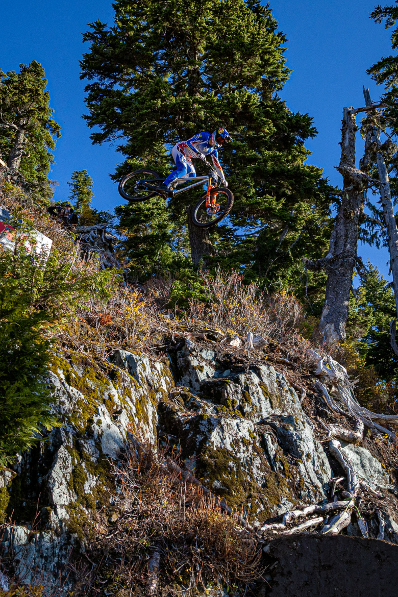 A mountain biker performing a jump off a rocky terrain, surrounded by trees under a clear blue sky. The rider is dressed in protective gear and colorful clothing, showcasing an action-packed moment in mountain biking.