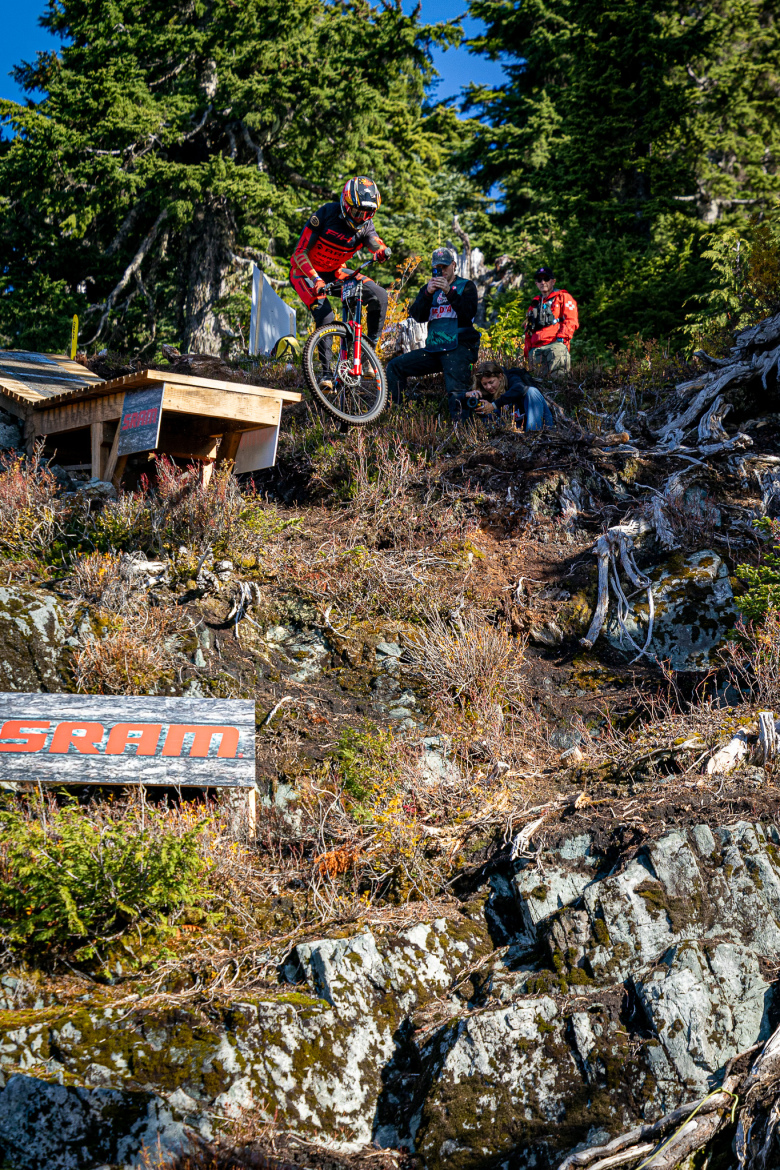 A mountain biker in red gear performs a jump off a wooden ramp in a natural outdoor setting, with rocky terrain and greenery in the background. Several spectators are watching and capturing the moment on camera. A SRAM sign is visible in the foreground.