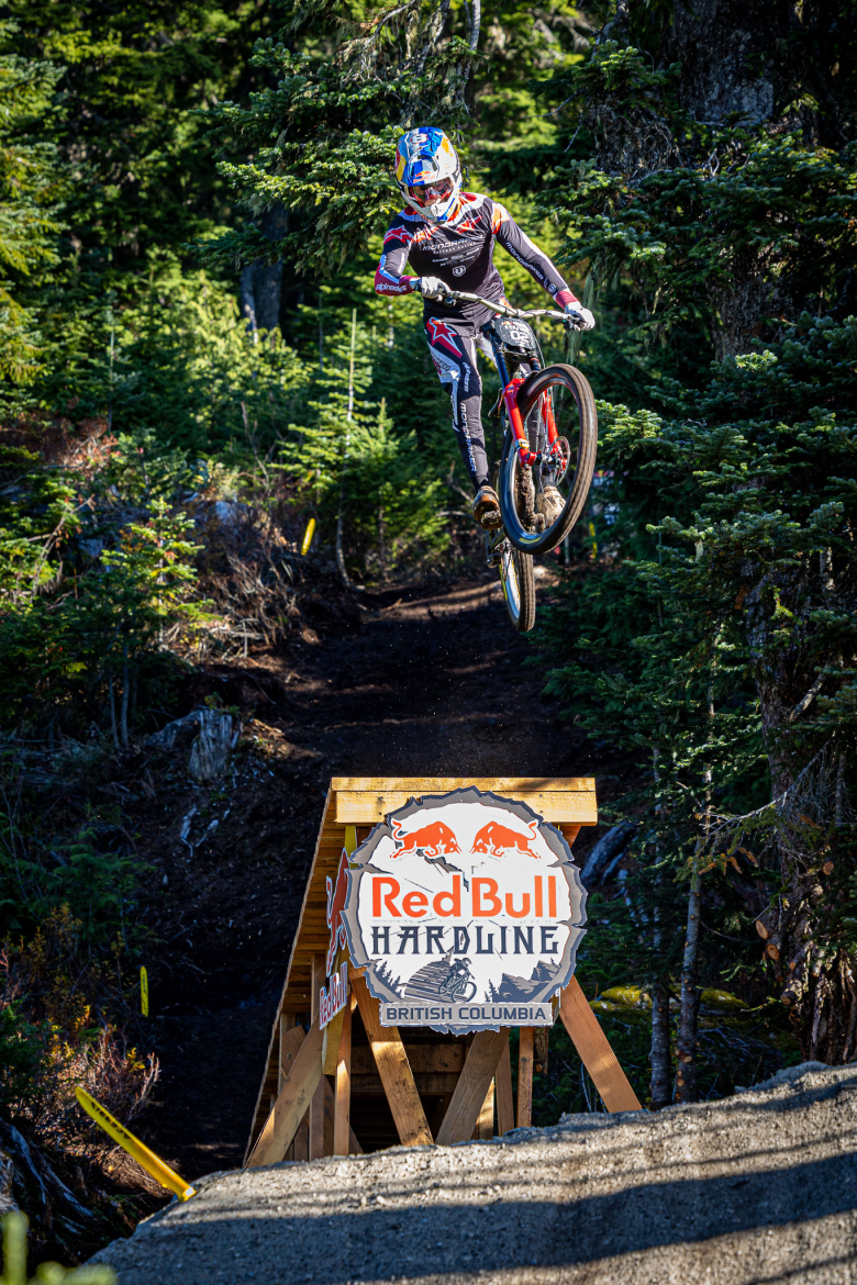 A cyclist performing a jump off a wooden ramp with a "Red Bull Hardline" sign, surrounded by trees in a forested area. The rider is in mid-air, showcasing their bike skills against a backdrop of rugged terrain in British Columbia.