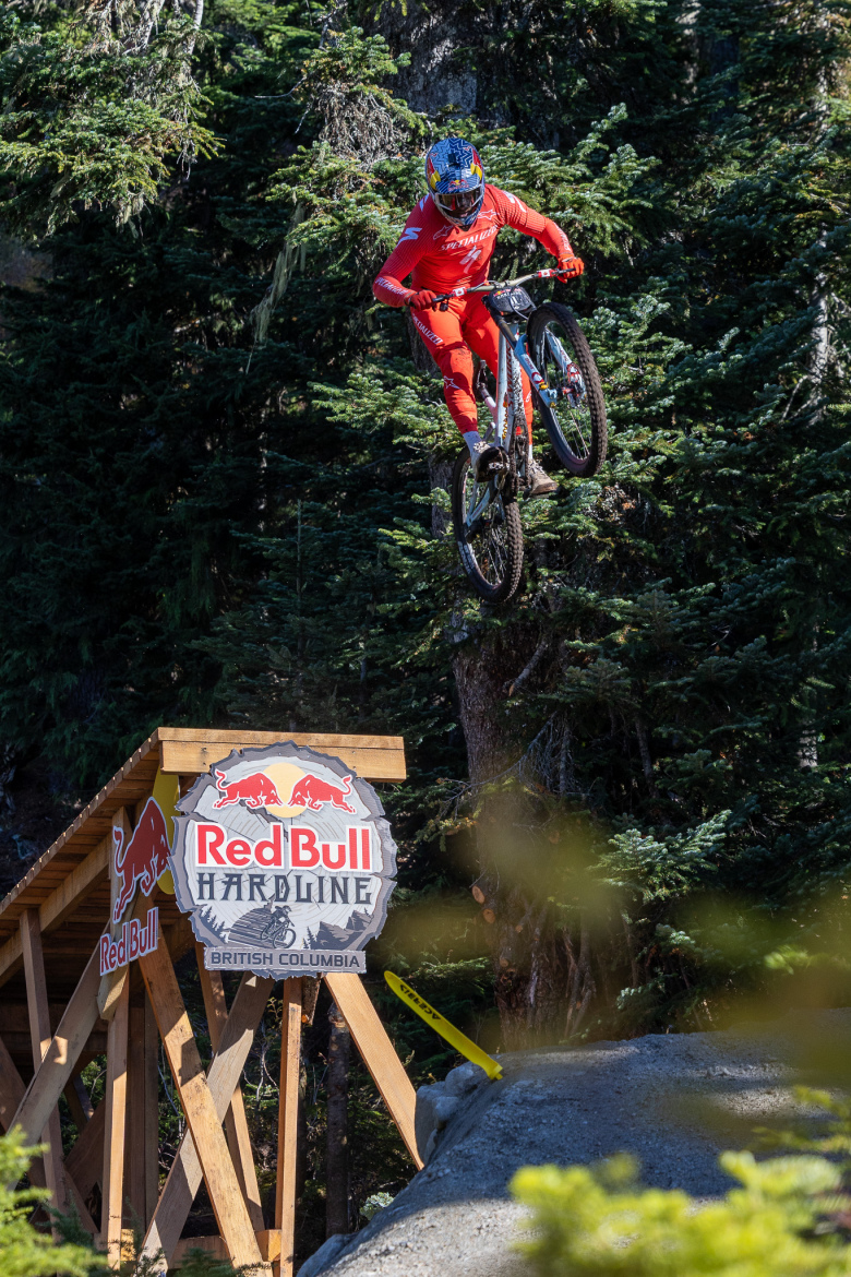 A mountain biker in a red outfit performs a jump over a wooden ramp with a "Red Bull Hardline" sign in the foreground. The scene is set in a mountainous area surrounded by lush green trees, showcasing an action-packed moment during the biking event.