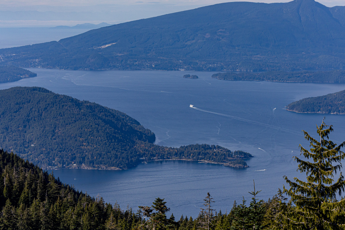 A panoramic view of a tranquil blue bay surrounded by forested hills, with a mountain in the background. A ferry is seen cruising through the water, leaving a wake behind. Several small islands can be spotted in the bay, with a clear sky above.