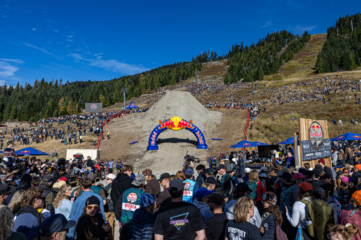 A large crowd gathers at an outdoor event, with a prominent dirt ramp featuring a colorful archway at the top. The background is filled with trees and a clear blue sky, and various tents and banners displaying branding can be seen among the spectators. The atmosphere is lively, with people dressed casually enjoying the event.