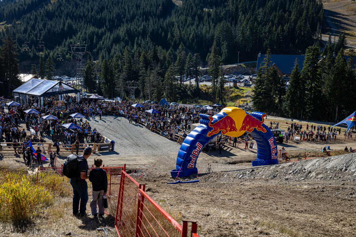 A bustling outdoor event scene at a mountain venue, featuring a large crowd gathered around a racecourse marked by a colorful inflatable Red Bull arch. In the foreground, a person and a child observe the festivities, while a clear blue sky and pine trees provide a scenic backdrop. Various tents and structures for vendors and participants are visible, along with additional spectators in the distance.