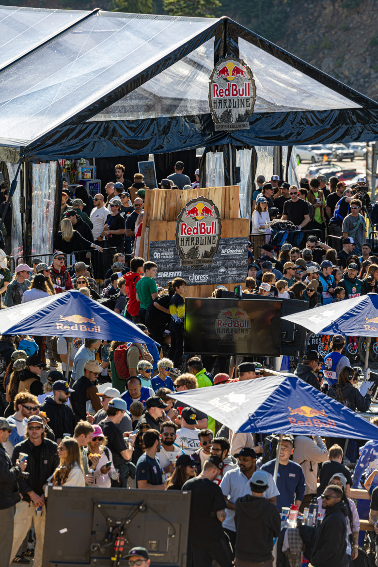 Crowd gathered at the Red Bull Hardline event in British Columbia, with tents and promotional banners visible. Attendees, wearing casual clothing and hats, are socializing, while a large screen displays the event logo. The atmosphere is lively, with a mix of excitement and anticipation.