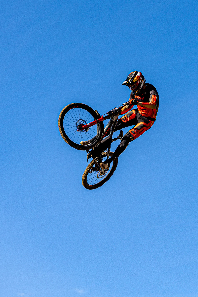 A mountain biker performing a mid-air trick against a clear blue sky. The rider is wearing a black and orange outfit, with a helmet and gloves, showcasing a dynamic pose while the bicycle is elevated off the ground.