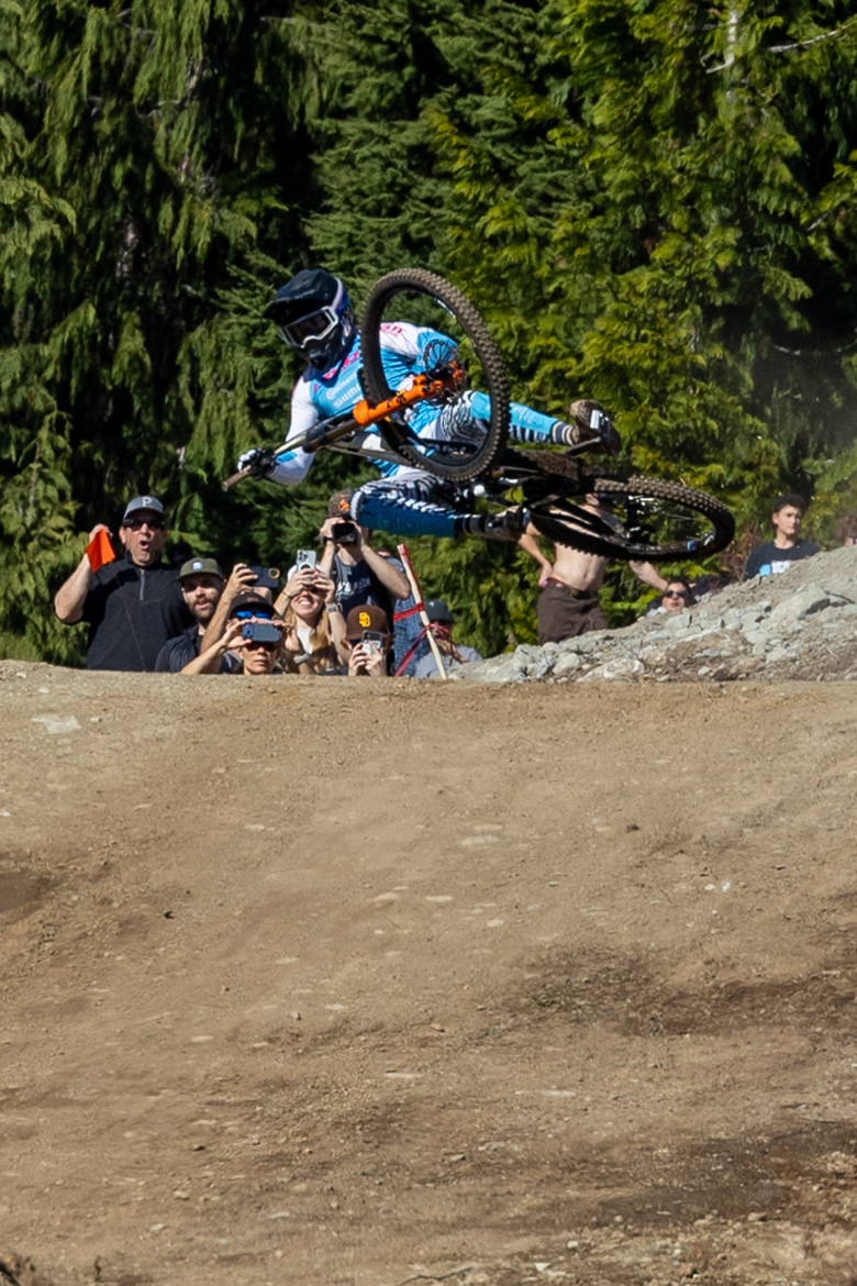 A mountain biker performing a jump on a dirt trail, surrounded by spectators who are taking photos and cheering. The background features lush green trees, and the biker is wearing a helmet and colorful gear.