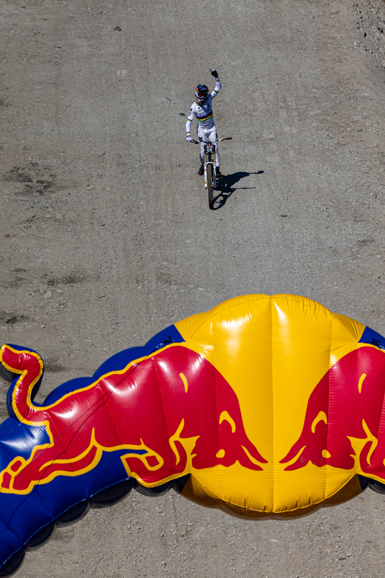 A cyclist in a racing outfit raises their hand in celebration as they ride past a large inflatable Red Bull arch on a dirt track. The image captures a sunny day with a clear view of the rider and the surrounding terrain.