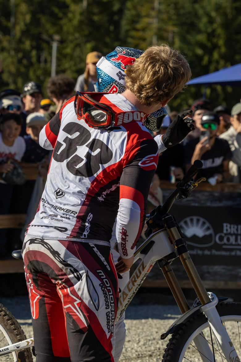 A mountain biker wearing a red and white jersey with the number 33 embraces another rider while standing next to their bike. In the background, a crowd watches the interaction with enthusiasm, surrounded by trees and outdoor event decorations.