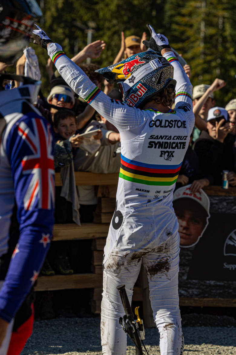 A mountain bike racer in a white racing uniform, showing a victory gesture to a cheering crowd, with muddy shorts indicating recent competition. The background includes fans enthusiastically displaying excitement, and a wooden fence lined with spectators.