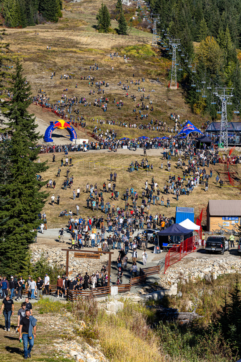 A large crowd of people gathered at Cypress Mountain for an outdoor event, with a backdrop of trees and ski lifts. The scene features a vibrant blue and red archway, tents, and a wooden sign indicating "Cypress Mountain." The atmosphere appears lively, showcasing attendees enjoying the day in a natural setting.