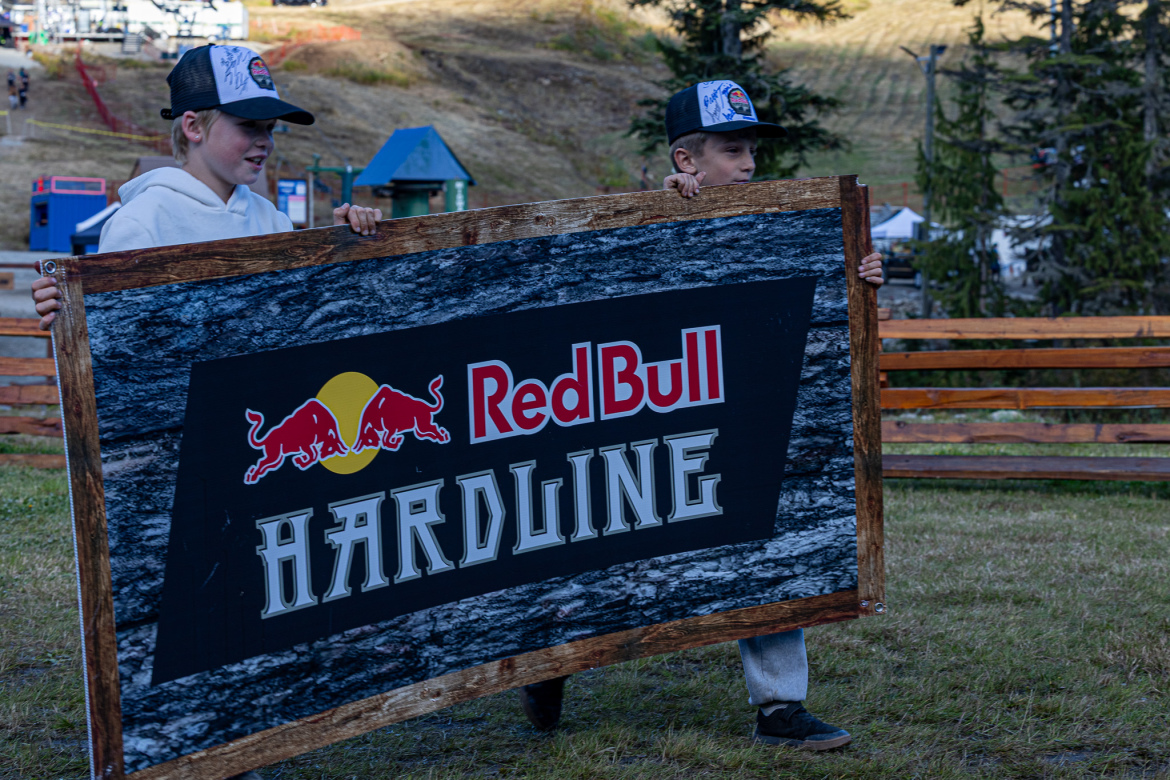 Two children are carrying a large sign that reads "Red Bull HARDLINE." They are outdoors, surrounded by trees and a grassy area, with a ski lift and equipment visible in the background. The children are wearing hats and casual clothing.