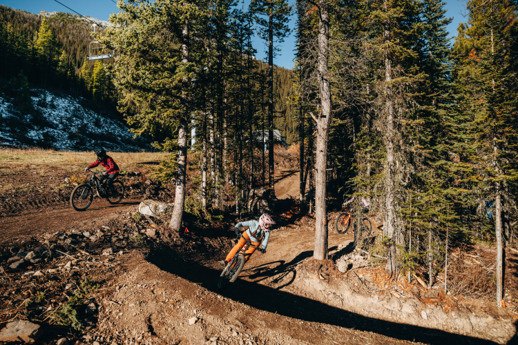 Mountain bikers navigating a winding dirt trail through a forested area, with tall pine trees and a chairlift visible in the background. One rider leans into a turn while another is seen in the distance. The scene is set against a backdrop of rugged terrain and clear blue skies.