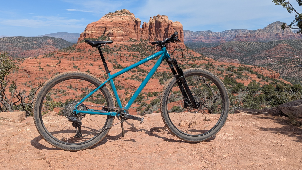 A mountain bike in a vibrant blue color, parked on a rocky terrain with red sandstone formations in the background. The landscape features a mix of greenery and distant mountains under a clear blue sky, highlighting an outdoor adventure setting.