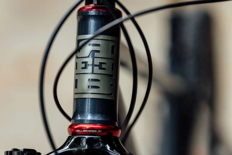 Close-up view of a bicycle's head tube featuring a metallic logo and a red headset cap, with cables positioned around it. The image highlights the craftsmanship and design details of the bike's components.