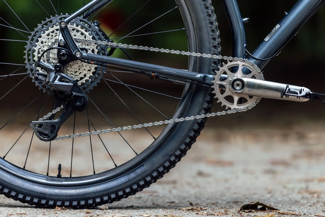 Close-up of a bicycle drivetrain, featuring a rear cassette, derailleur, and chain on a mountain bike tire resting on a gravel surface.