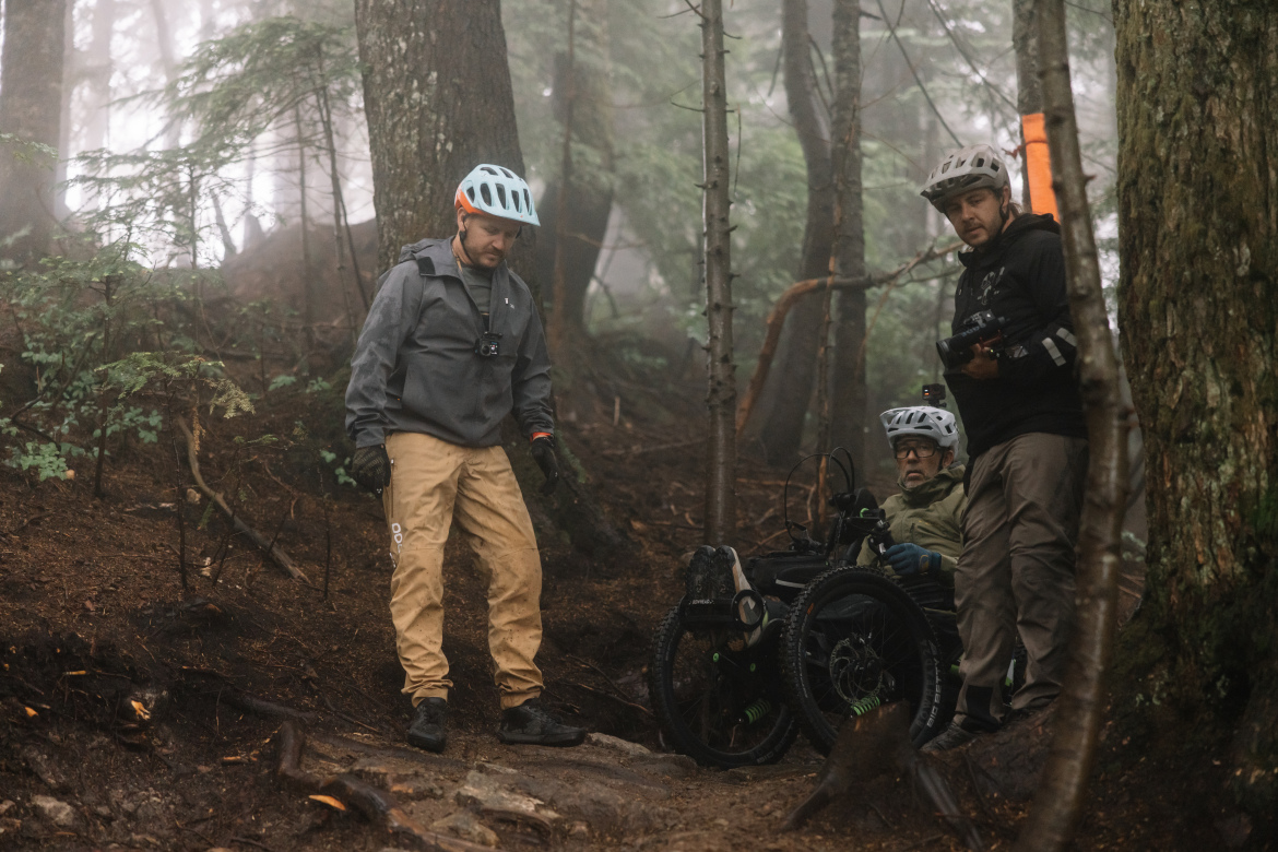 Three individuals in a forested area are working together in a misty environment. One person is wearing a helmet and looking down, while another is crouched beside a specialized bike with three wheels, indicating a focus on adaptive cycling. The third person stands nearby with a camera, capturing the scene. The ground is uneven and covered with leaves and roots, suggesting a rugged terrain.