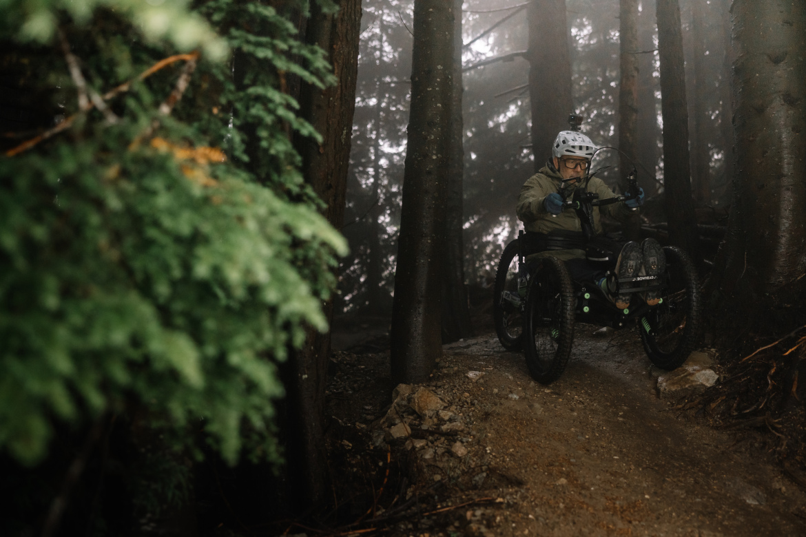 A person in an all-terrain wheelchair navigates a rugged trail through a misty forest, surrounded by tall trees and greenery. The individual wears a helmet and goggles, showcasing determination while maneuvering through the challenging terrain.
