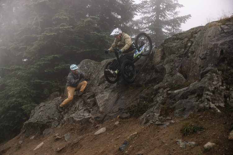 A mountain biker navigates a rocky descent while a second person watches and encourages from a lower position. The scene is set in a foggy forest, adding a mysterious atmosphere to the challenging terrain.
