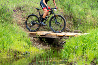A cyclist in red and black apparel rides across a wooden bridge over a small pond, surrounded by lush green grass and trees. The cyclist's reflection is visible in the water.