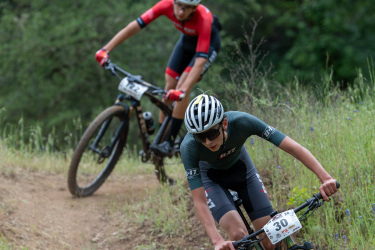 Two mountain bikers compete on a dirt trail surrounded by greenery. The rider in the foreground, wearing a dark jersey and helmet, leans forward as they navigate the curve of the trail. The second rider trails behind, dressed in a red jersey and black shorts, maintaining a focused posture. The scene captures the intensity and excitement of a mountain biking race.