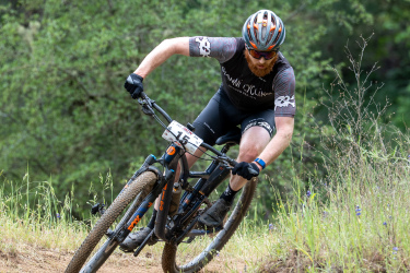 A male mountain biker wearing a helmet and cycling gear leans into a turn on a dirt trail surrounded by greenery. The cyclist is focused on navigating the terrain, with dirt and grass in the foreground and blurred trees in the background.