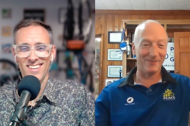 Two men are participating in a video call. The man on the left has short hair, glasses, and is wearing a patterned shirt. He is smiling and speaking into a microphone. The man on the right has short, light hair and is wearing a blue shirt with a logo. He is also smiling and gesturing. In the background, there are various awards and biking equipment.