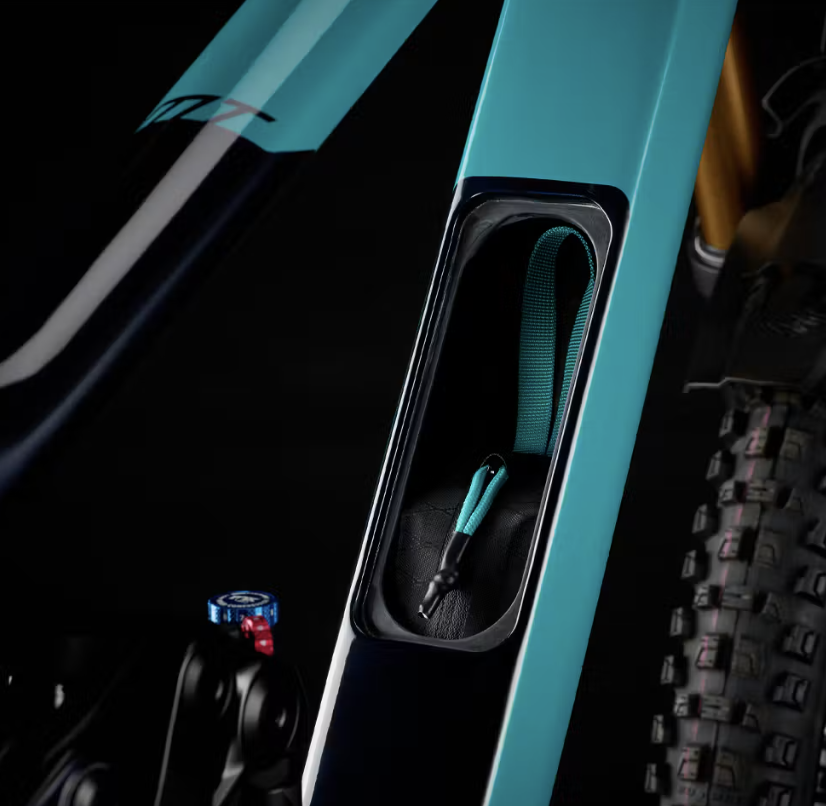 A close-up view of a bicycle frame featuring a turquoise and black color scheme. The image highlights a storage compartment in the frame, which contains a small black and turquoise item. In the background, part of the bike's suspension system and a textured tire are visible. The overall composition is set against a dark backdrop.