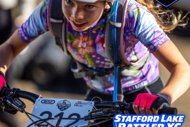 A young girl competes in a mountain biking event, wearing a white helmet and a colorful floral jersey. She appears focused as she grips the handlebars of her bike, which has a race number pinned to it. The background shows a blurred crowd, suggesting a lively outdoor event. Text overlay includes details about the "Stafford Lake Rattler XC" and a juniors fundraiser, with the date November 8, 2026.