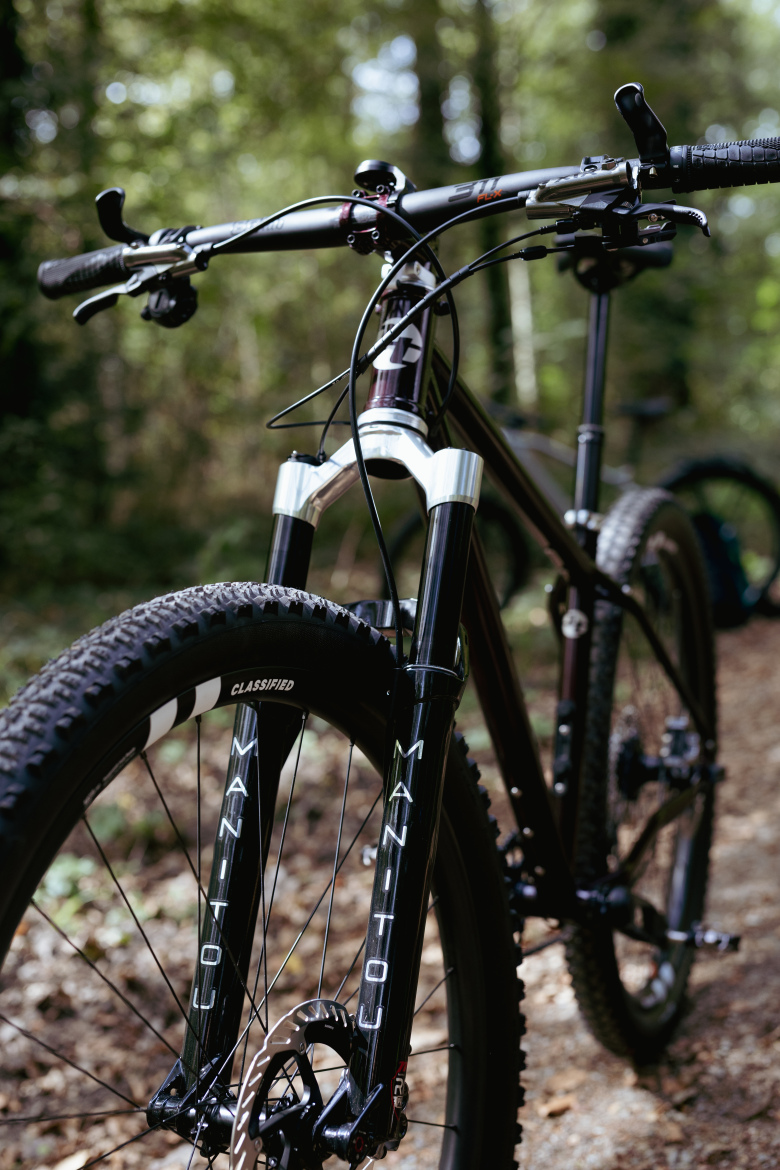 Mountain bike parked on a forest trail, showcasing the front fork and handlebars. The bike features tubeless tires and detailed components, with a blurred green background of trees and nature.