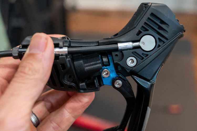 A close-up image of a hand holding a black mechanical component, showcasing intricate details such as metal springs and a blue part, typically found in a bicycle's shifting mechanism. The background is slightly blurred, with hints of a workshop environment.