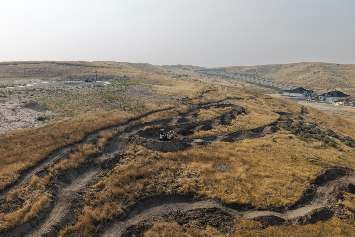 Aerial view of a dry, hilly landscape featuring construction activity. A machine is digging in a winding, dirt path amidst tall, dry grass. In the background, new residential buildings and a roadway are visible. The sky is hazy, suggesting a lack of visibility.