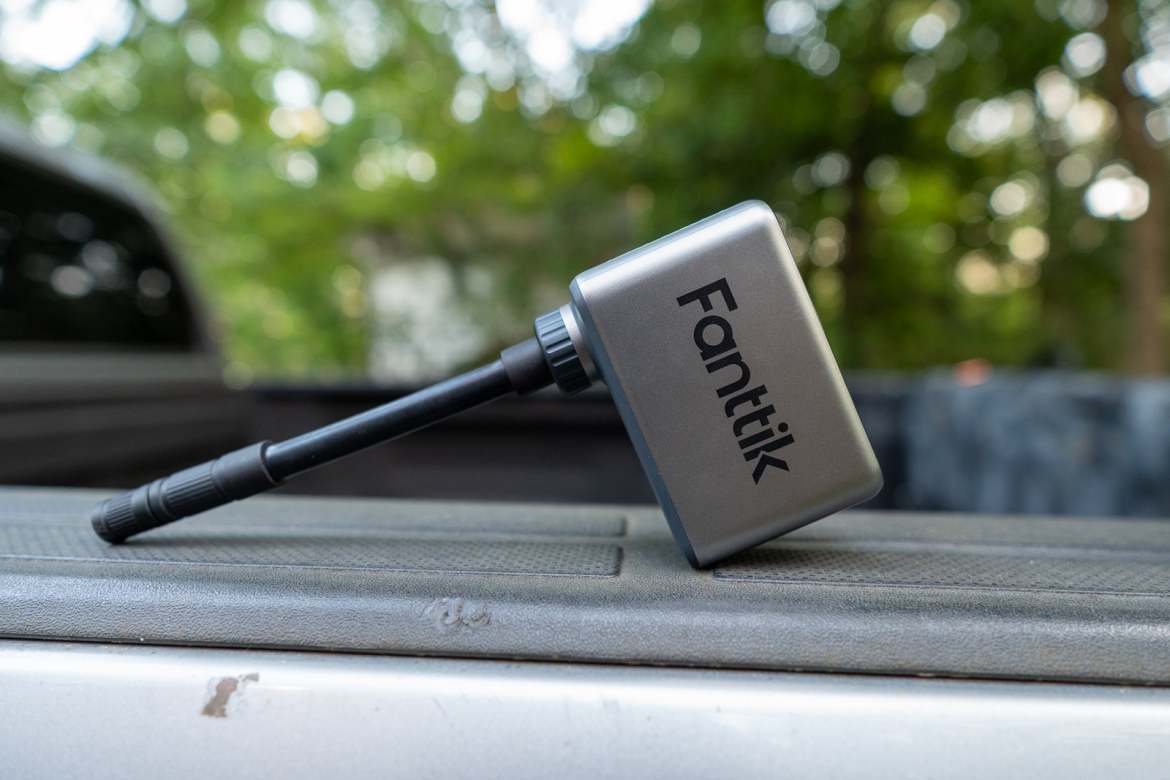 A compact Fanttik device with an antenna is resting on a textured surface in an outdoor setting, surrounded by blurred greenery. The device features a sleek, metallic design with the name "Fanttik" prominently displayed on its side.