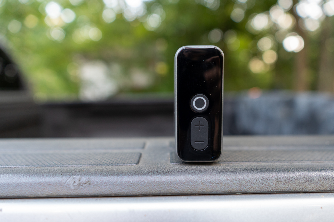 A black Bluetooth speaker with a circular power button and volume controls sits on the surface of a truck bed, with a blurred green background.
