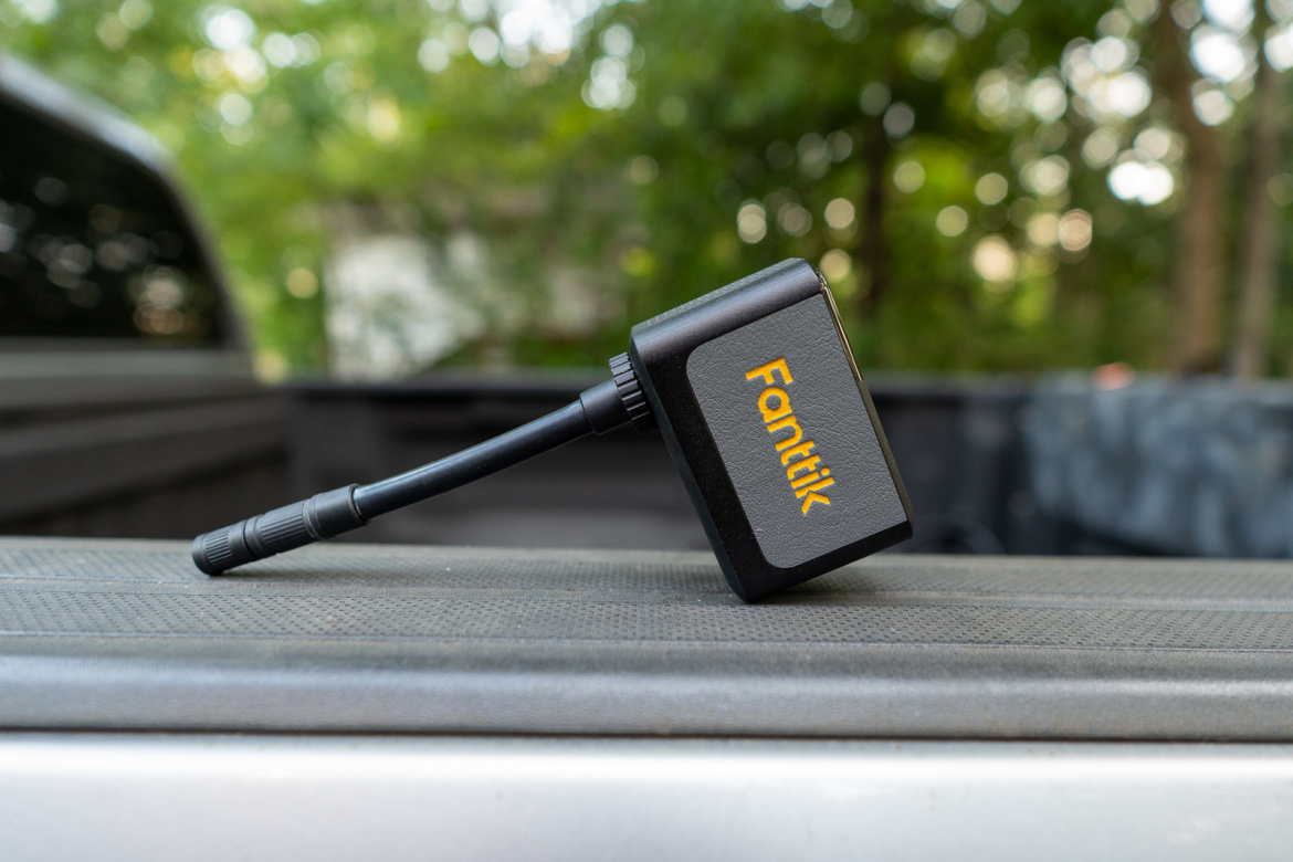 A compact Fanttik device with a black body and an orange logo, resting on a textured surface in a natural outdoor setting. The background features soft, blurred greenery, suggesting a casual environment, possibly on the bed of a truck.