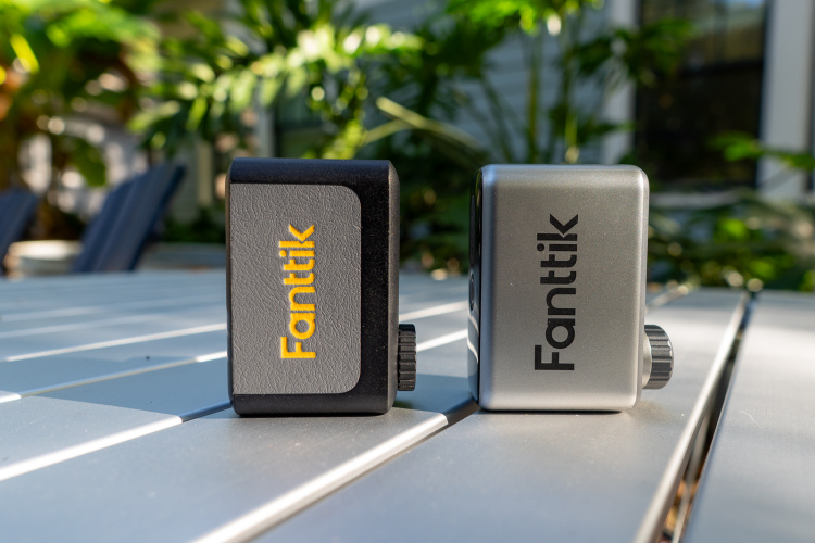 Two devices labeled "Fanttik" are displayed side by side on a light-colored table. The one on the left is primarily black with yellow lettering, while the one on the right is silver with black lettering. In the background, there are hints of lush green plants, creating a vibrant outdoor setting.