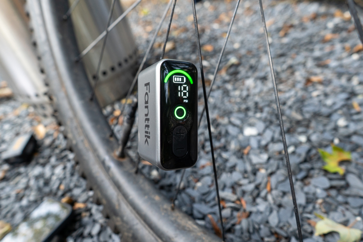 A close-up of a Fanttik digital tire pressure monitor attached to a bicycle wheel, displaying a pressure reading of 18 PSI on its screen. The background features gravel and a blurred metallic surface.