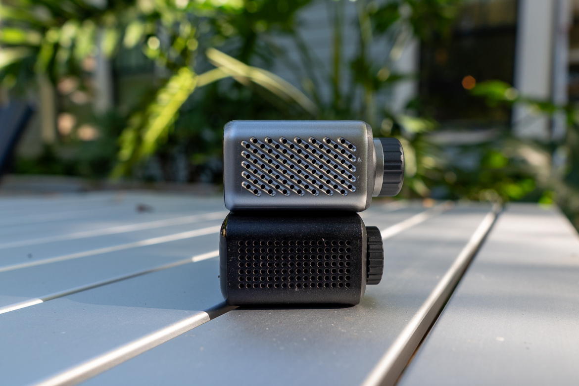 A close-up image of two compact audio devices, one gray and one black, stacked on top of each other. The devices feature perforated designs on their sides and a textured knob for adjusting settings. The background consists of green foliage, while the foreground shows a light-colored wooden surface.