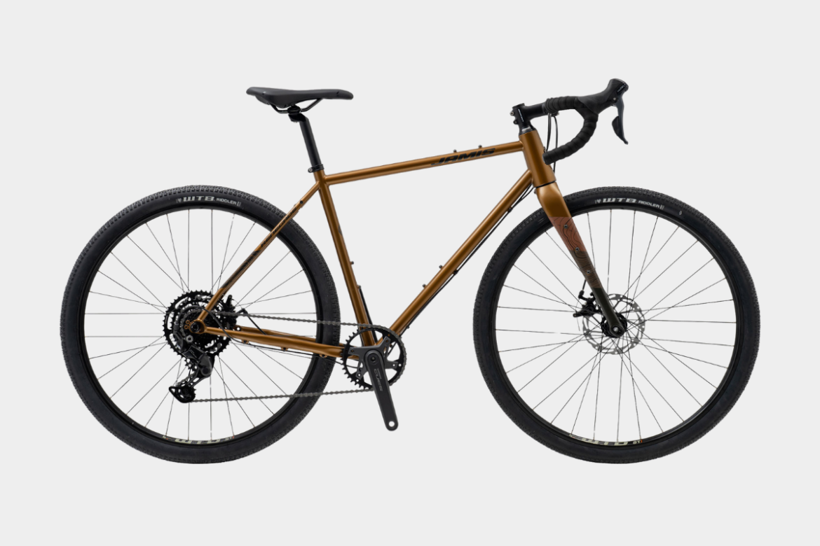 A modern gravel bike featuring a sleek, bronze frame, drop handlebars, and thick tires designed for off-road terrain. The bike is shown from the side, highlighting its disc brakes and unique design elements, suitable for versatile cycling adventures.