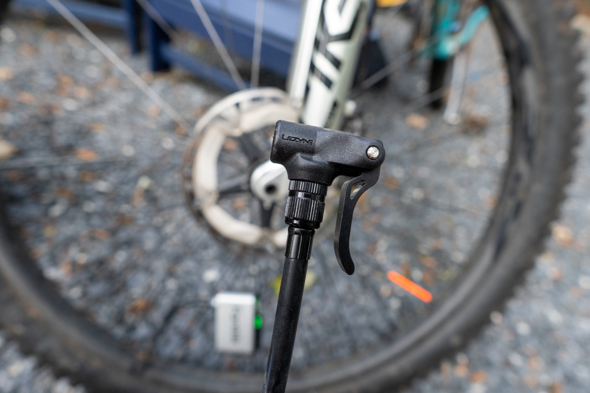 Close-up of a bicycle tire inflator valve, featuring a black nozzle with a lever mechanism, positioned in front of a bicycle wheel resting on gravel. The wheel's rim and brake rotor are faintly visible in the background.