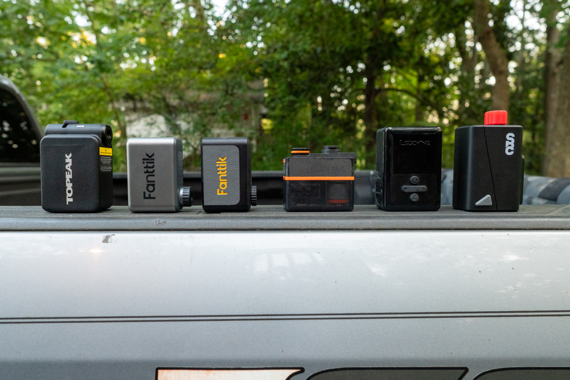 A lineup of six portable devices for cycling and outdoor activities, displayed on the edge of a truck bed. The items vary in design and color, showcasing brands like Topeak and Fanttik, with the background featuring soft-focus greenery.