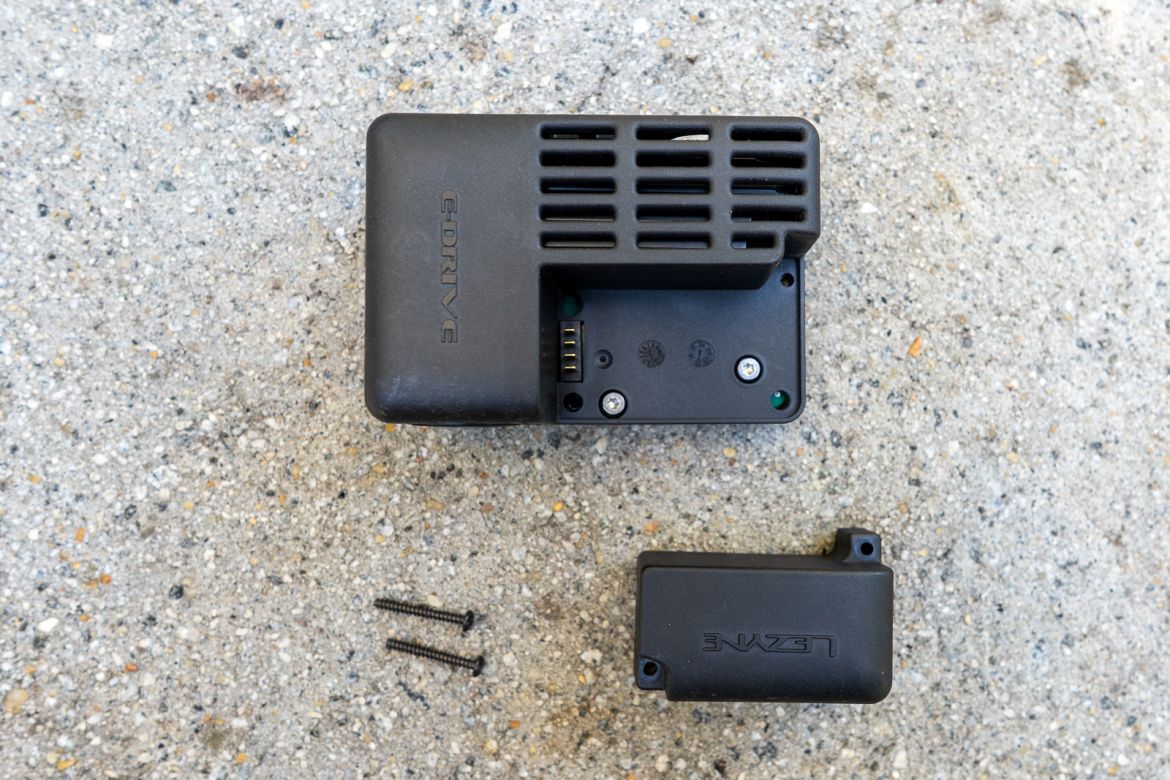 A black electronic device labeled "E.DRIVE" placed on a textured surface, accompanied by a small black component and two screws. The device features a vented design with visible ports and screws on its underside.