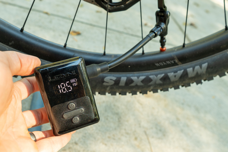 A hand holding a digital tire pressure gauge, displaying a reading of 16.5 PSI, while measuring the pressure of a bicycle tire. The gauge is connected to the tire's valve, with a close-up view of the wheel and tire labeled "MAXXIS."