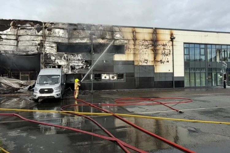 Firefighters extinguish a large commercial building that has sustained severe fire damage, with charred walls and blackened windows. A fire truck is parked nearby, and hoses are laid out on the ground as water is sprayed on the affected area. The scene is cloudy and wet, indicating recent firefighting activities.