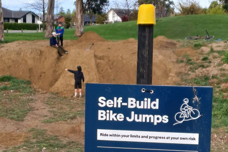 Three children are seen at a dirt bike jump site, with one child sitting atop a mound, another digging, and a third pointing. A sign nearby reads "Self-Build Bike Jumps" and advises riders to "Ride within your limits and progress at your own risk." The background features trees and houses.
