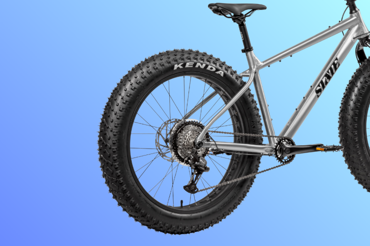 A close-up view of a silver fat bike, featuring large, textured tires with "Kenda" branding. The bike's frame is prominently displayed, showcasing sleek lines against a gradient blue background. The rear wheel, drivetrain, and components are visible, emphasizing the bike's rugged design for off-road use.